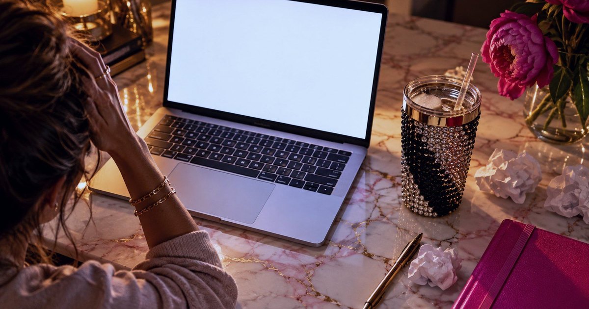 A woman at her laptop in the evening, head in her hands, with a blank screen and crumpled paper on a marble desk